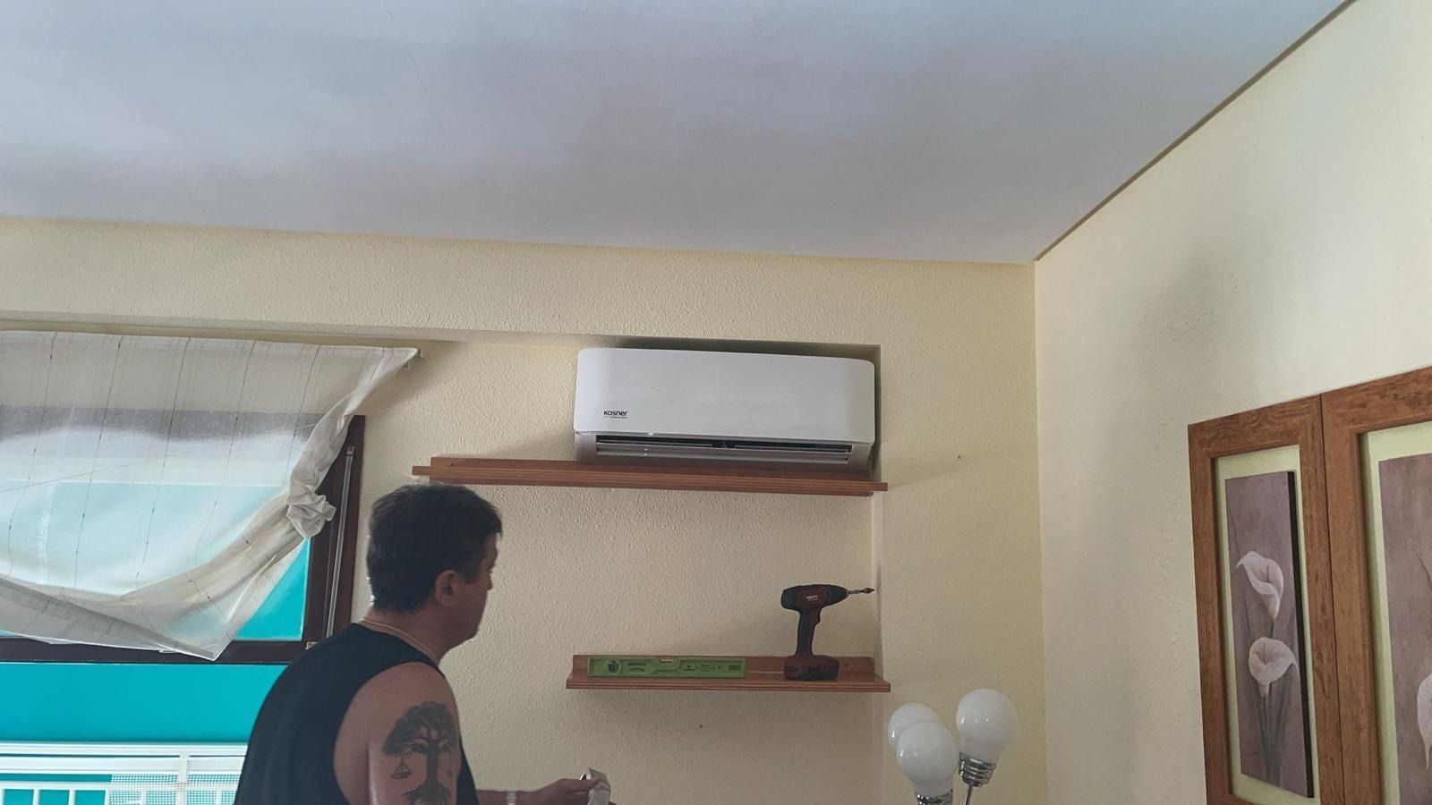 Un hombre con un tatuaje en el brazo está de pie en una sala de estar con un aire acondicionado en la pared.